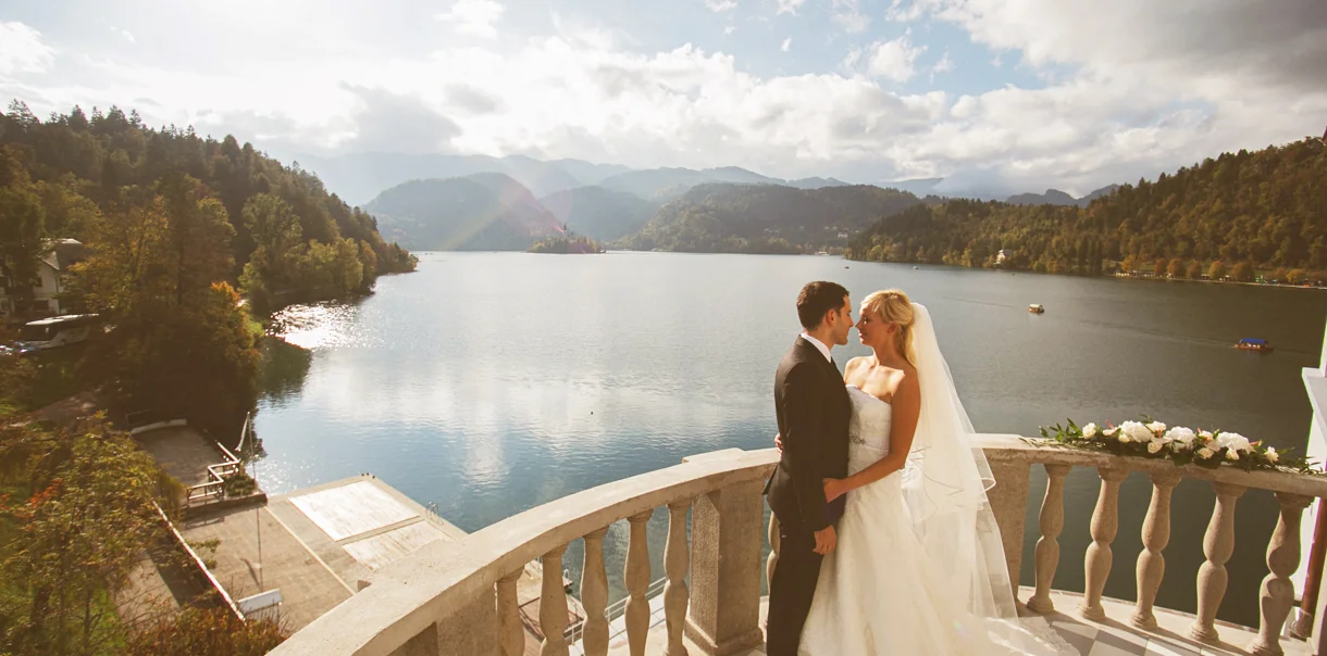 Novoporočenca se v poročnih obleki objemata in zaljubljeno gledata na terasi ob jezeru