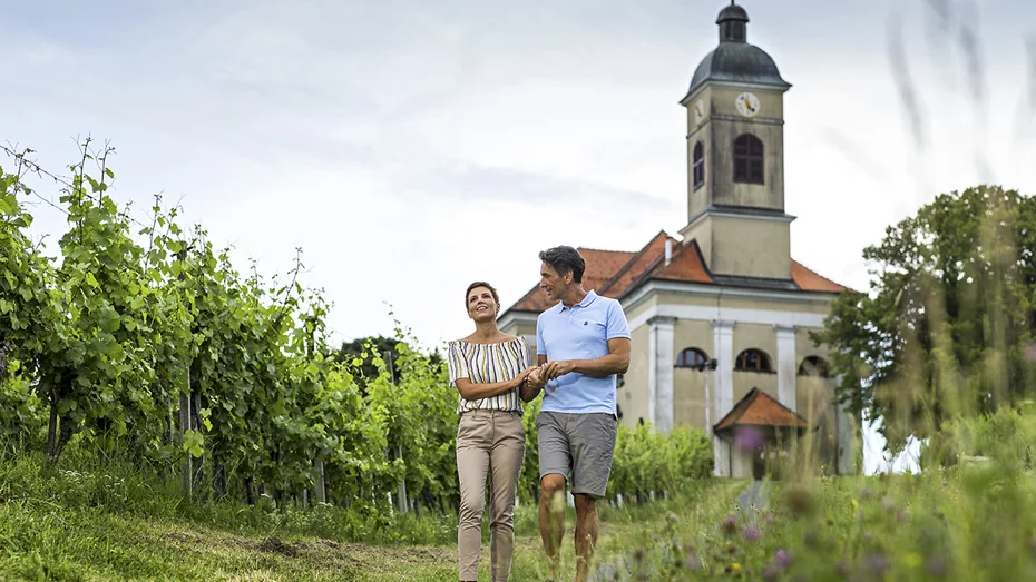 Moški in ženska se v športnih oblačilih sprehajata po potki iz cerkve med vinogradi
