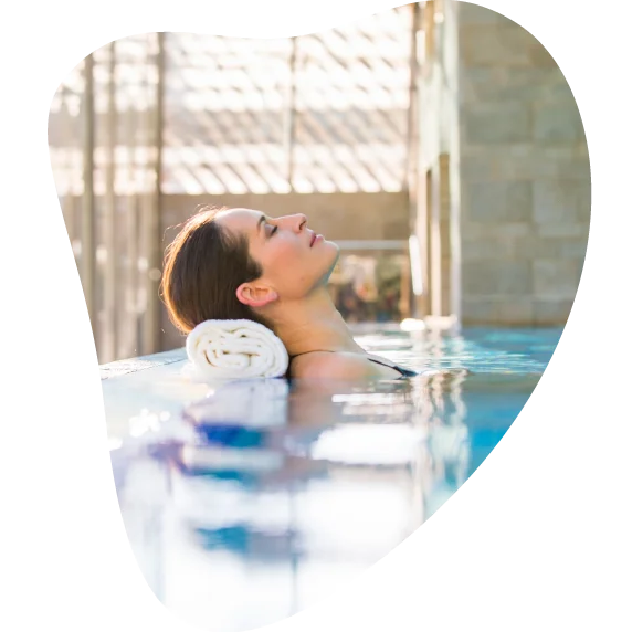 Ženska z zaprtimi očmi je naslonjena na belo brisačo in sproščeno uživa ob robu bazena