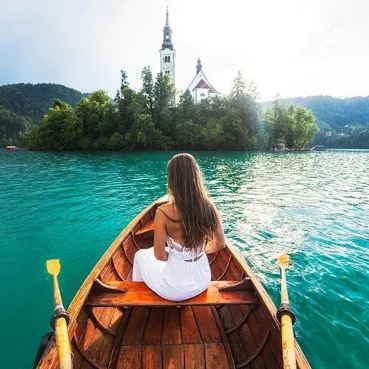 Ženska v beli obleki sedi na čolnu na blejskem jezeru in gleda cerkev na otoku