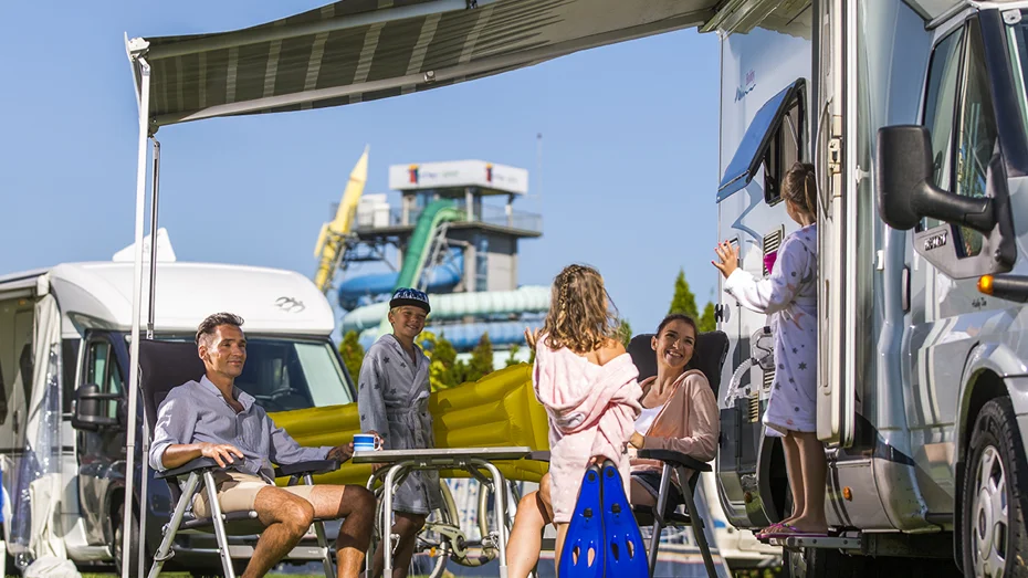 Pet ljudi stoji in sedi za mizo pod baldehinom avtodoma in se pogovarja