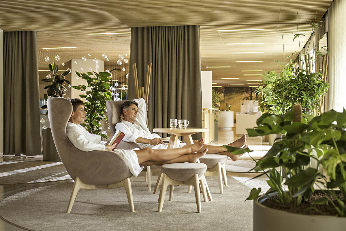 Moški in ženska v belih kopalnih plaščih počivata na ležalniku v zelenem sproščujočem prostoru s knjigo v roki