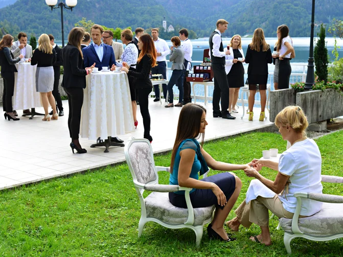 Skupina ljudi v poslovnih oblekah se druži ob visokih mizah na terasi pri jezeru