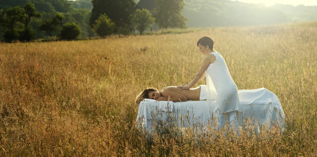 Maserka v beli obleki masira žensko, ki leži na masažni mizi na travniku