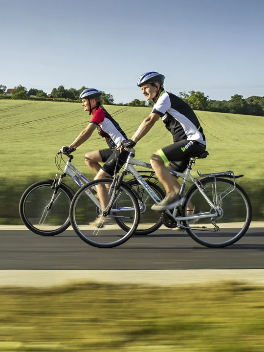 Dva kolesarja v kolesarski opremi kolesarita drug ob drugem na kolesarski stezi ob zelenih travnatih površinah