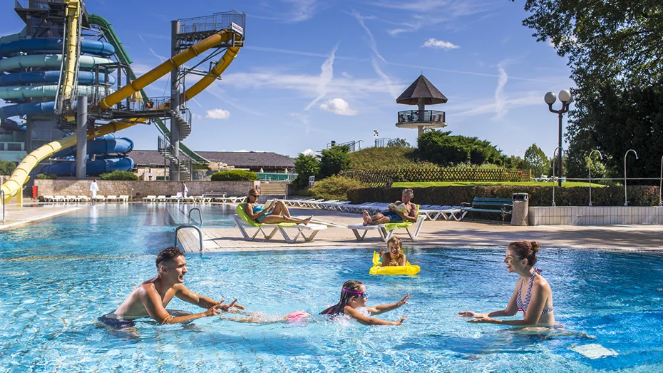 Mlajša deklica se v zunanjem bazenu uči plavati, oče in mama pa ji pri tem pomagata