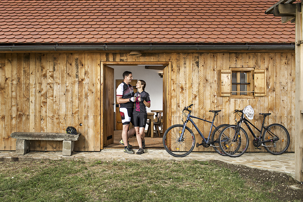 Mlad kolesarski par, oblečen v kolesarska oblačila, stoji pred leseno hišico in se pogovarja